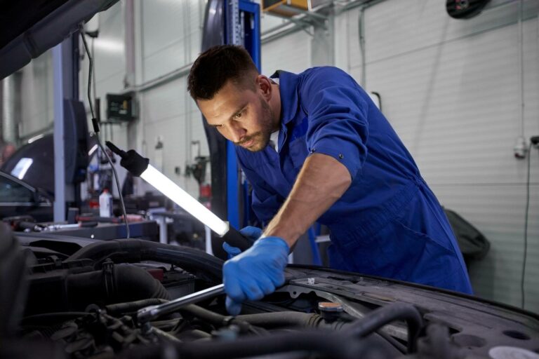 Mechanik w niebieskim kombinezonie i rękawiczkach pracuje przy silniku samochodowym, używając klucza. Fluorescencyjne światło robocze oświetla obszar. Garaż jest dobrze oświetlony i uporządkowany, w tle widać narzędzia i sprzęt samochodowy.